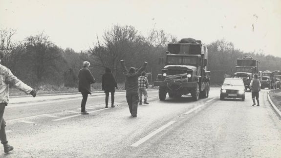 A group of people standing on a road, intercepting a convoy of large trucks. Some individuals have their arms raised.