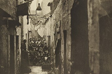 An old black and white photograph people in an alley between tenements. There is washing hanging above them.