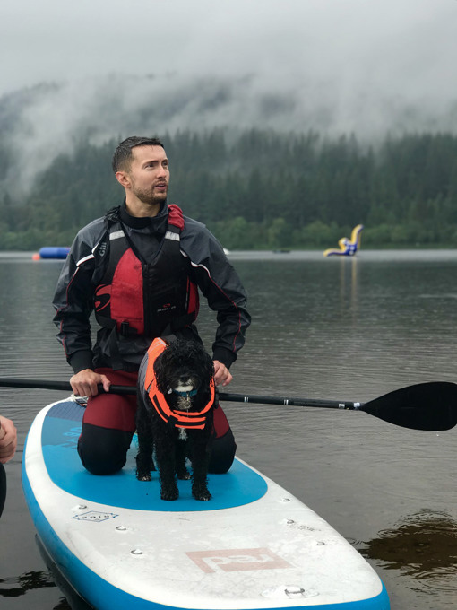 Person kneeling on a paddleboard with a black dog in a life jacket on a misty loch, with forested hills in the background.