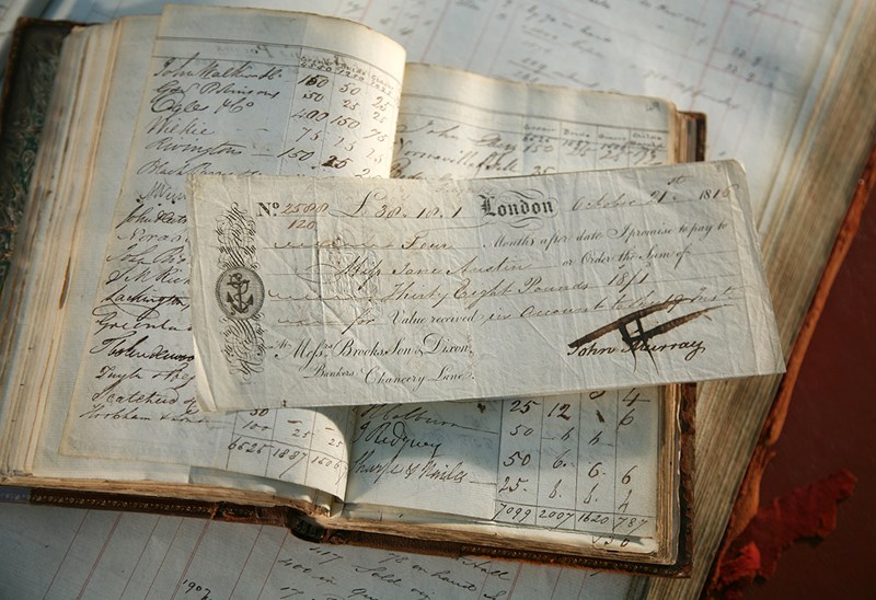 A smaller book lying open on top of another larger book. They both are full of handwritten notes. There is an old cheque lying on top.