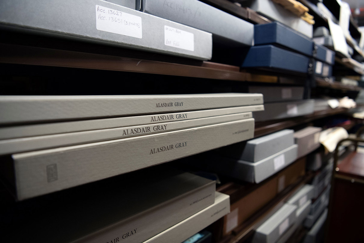 Thin boxes of archives sitting on shelves in the stacks. They have 'Alasdair Gray' written on the spines of the boxes.