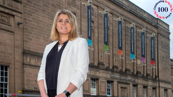 Amina Shah standing in front of the National Library of Scotland. In the corner is the Library's centenary logo.