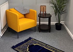 A photo of a room. There is an armchair, with a small table, lamp, plant, and prayer rug on the floor.