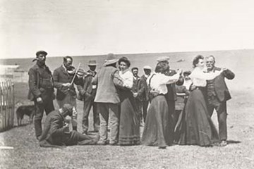 A black and white photo of men and women in old dress dancing and playing instruments outside.