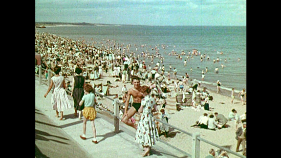 Film still showing a man talking to a woman next to a crowded beach.