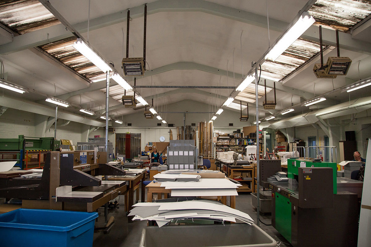 A view inside a large box workshop. There are lots of tables with sheets of cardboard on top of them and big machines to make the boxes.