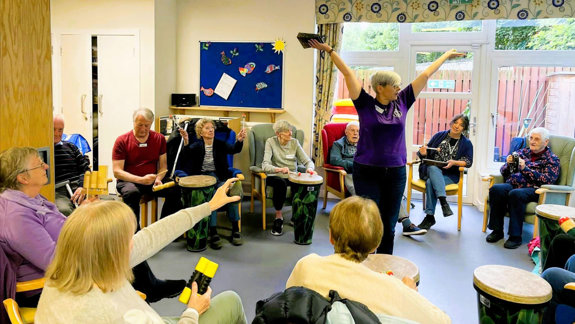 A women stands in the centre of a room surrounded by seated people using musical instruments.