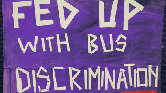 A close-up of a protest sign reading ‘FED UP WITH BUS DISCRIMINATION’ in white text on a purple background.