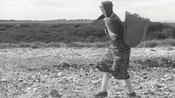 Black and white still of a film showing a person walking on rocky terrain carrying a large basket on their back. They are wearing a patterned dress and a hat. The background shows an expansive landscape with low vegetation, distant hills, and a cloudy sky.