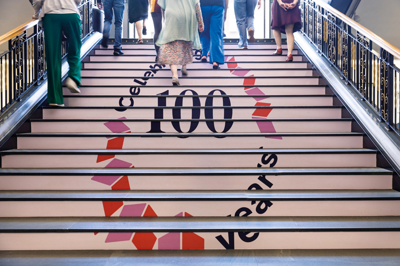 A group of people walking up the main stairs in the Library which have the centenary logo on them.