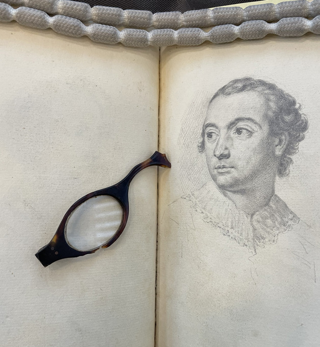 An open book displaying a detailed line drawing of a person on one page, with a small handheld magnifying lens resting across the opposite page.