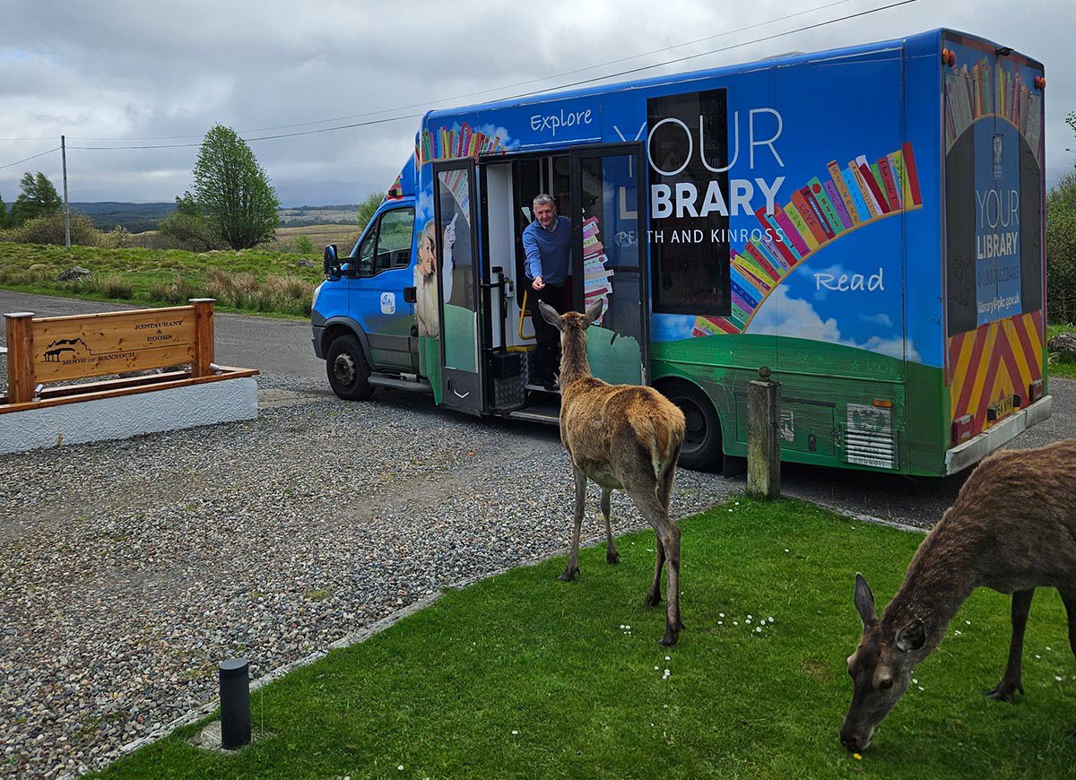 A mobile library with the librarian hanging out the door looking at two deer.