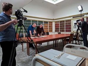 In the background there are two men standing over a table looking at some collection material. In the foreground, two other men are filming them.