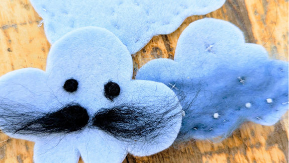 Three felt clouds on a table, one has eyes and a moustache.