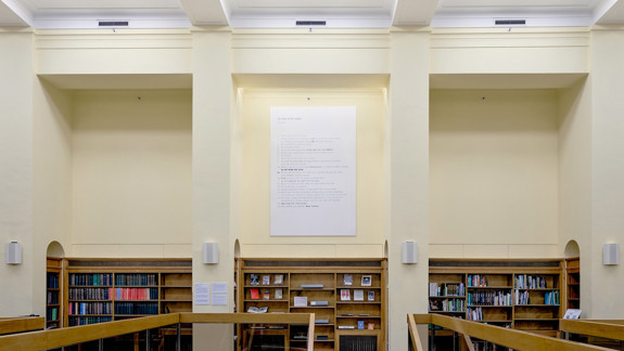 Large text-based artwork mounted high on the wall of a library reading room, above wooden bookshelves and study desks, framed by tall cream-coloured columns and a high ceiling.