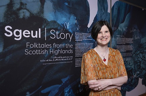 Photo of Kirsty MacDonald standing in front of a panel for an exhibition which reads "Sgeul | Story. Folktales from the Scottish Highlands".