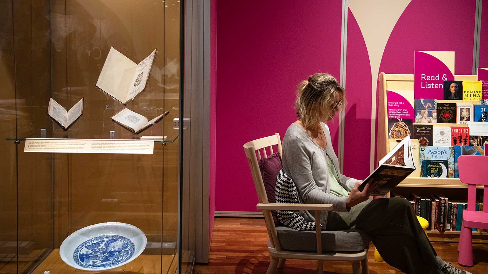 A woman sits in a chair reading next to a display case of books