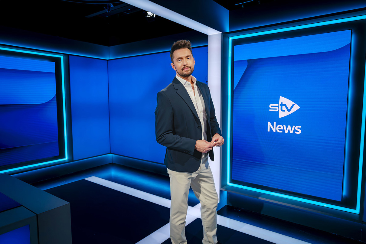 Person standing in a television studio beside a screen displaying the STV Weather logo.