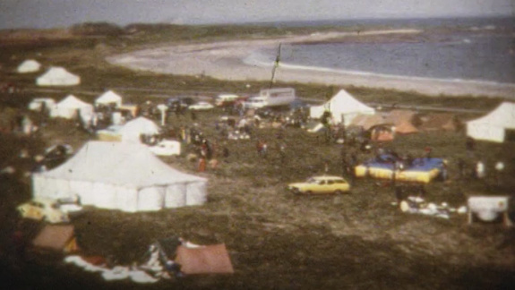 Still of a video showing a campsite near a beach with several tents and vehicles. People are gathered and engaged in various activities.