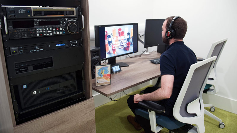 Man sits at a desk watching a film on the screen. To his left, a large technology stack sits in a cabinet.