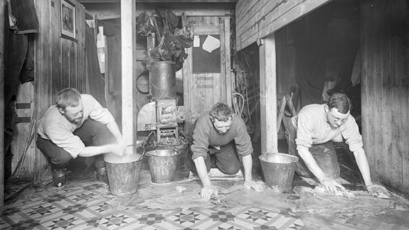 A monochrome image of three men on their hands and knees scrubbing a tiled floor of a ship.