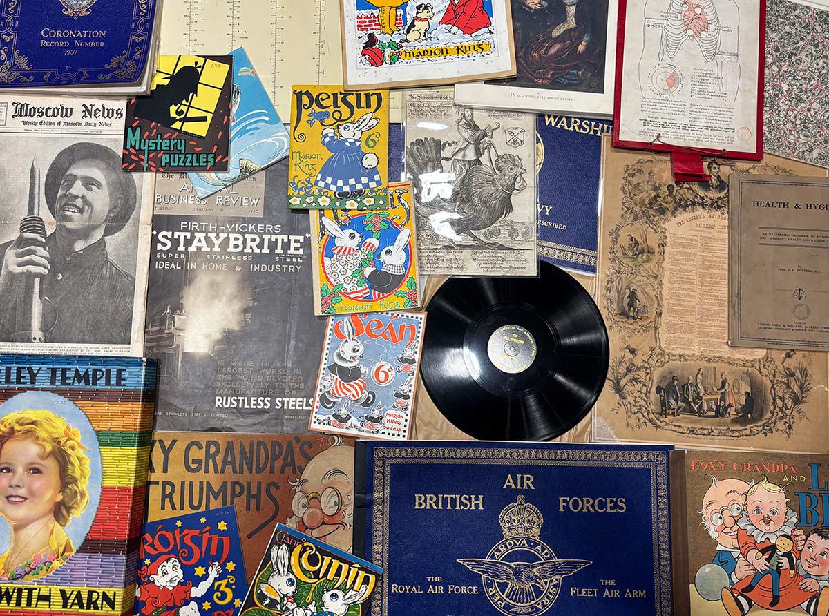 A collection of vintage items including comic books ("Pip," "Roisin," "Percy"), a vinyl record, a Shirley Temple book, "British Air Forces," and a "Moscow News" newspaper, all arranged together with other printed materials.