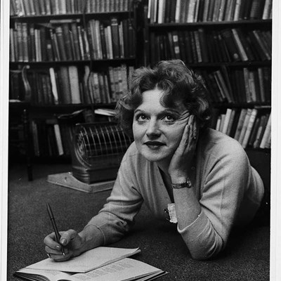 A photograph of Muriel Spark lying on the floor writing in a notebook.