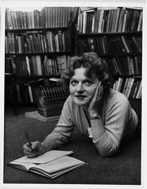 A photograph of Muriel Spark lying on the floor writing in a notebook.
