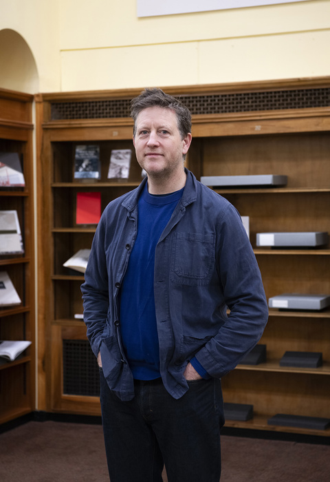 A person standing in a library reading room, with wooden bookshelves and display cases behind them.Peter 