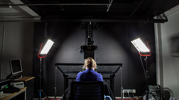 Person seated at a digitisation workstation beneath studio lights and imaging equipment.