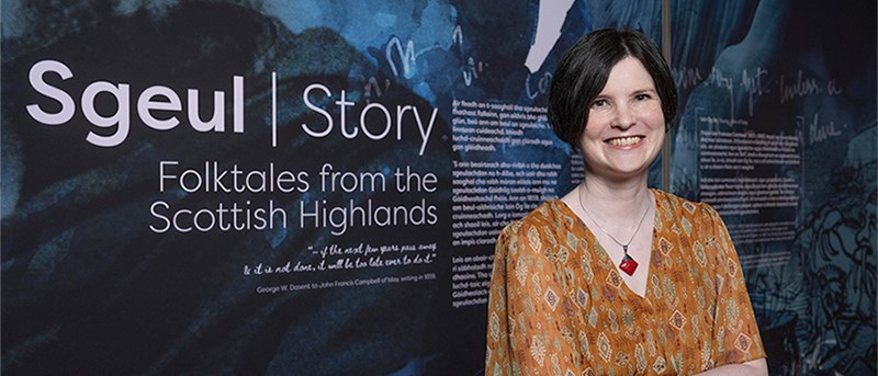 Photo of Kirsty MacDonald standing in front of a panel for an exhibition which reads "Sgeul | Story. Folktales from the Scottish Highlands".