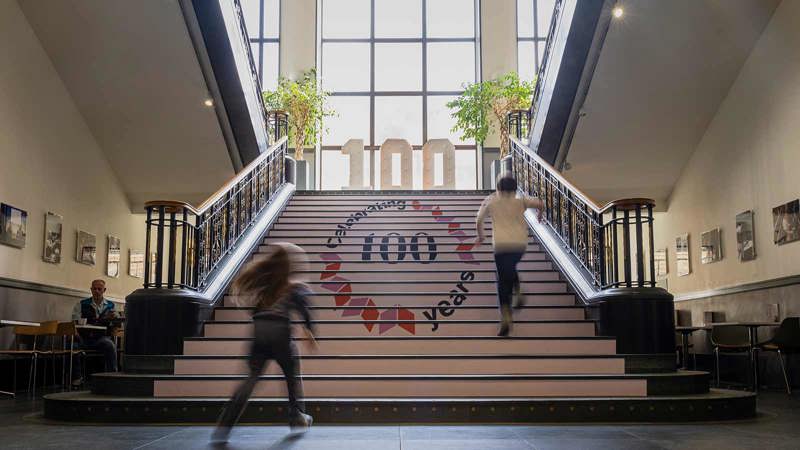 Staircase with 'Celebrating 100 years' on the steps, two people ascending, and a large '100' sign with plants at the top.