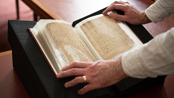 Hands turning pages of an open historic manuscript displayed on a book cradle.