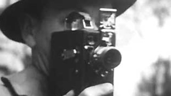 Black and white still of a man holding a super-8 camera up to his face.