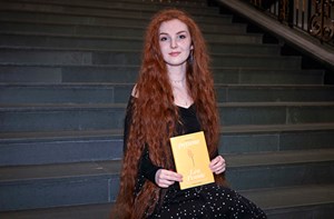 A woman with long red hair sitting on the steps of the library. She is holding a book entitled "poyems" by Len Pennie.