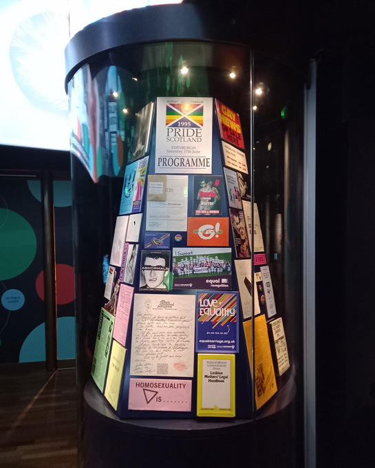 A circular exhibition case full of LGTQT+ archival material and ephemera.