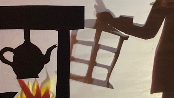 A cut out of a pot hanging over a fire and in the background a shadow of a person holding a lantern.