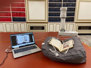 A visualiser set up. There is a book sitting on a book cushion on the table. Above it is the visualiser which is sending a live birds eye image of the book to the laptop.