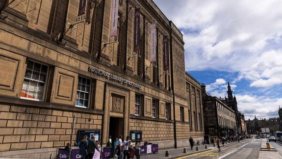 Exterior of the National Library of Scotland in Edinburgh, a large stone building with tall windows and banners, situated on a busy street with pedestrians. 