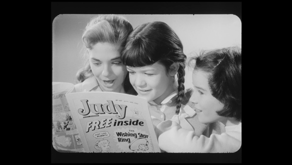 Black and white still image from advert for Judy magazine. Three girls crowd around an issue of the magazine.